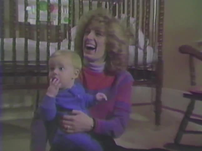 A loving mother laughs while holding her baby boy on the nursery floor in a sweet vintage home movie