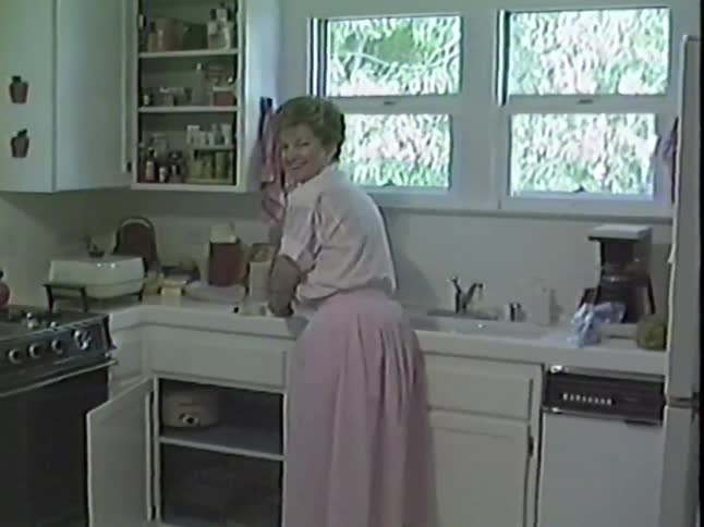 Two women smile and wave for the camera in a retro 1980s kitchen in this vintage home video
