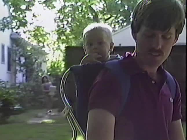 A vintage home video shows a smiling father carrying his baby in a backpack carrier through a sunny suburban yard