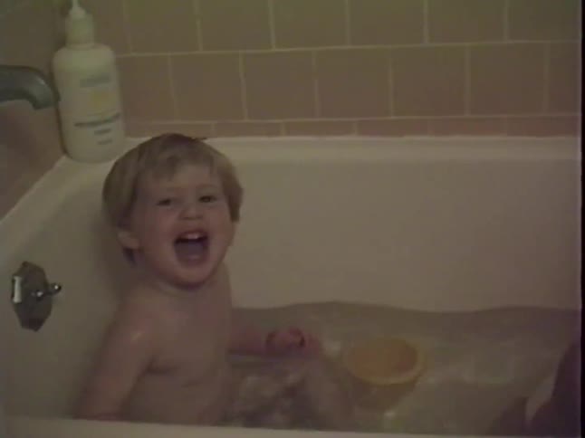Two young children splash and play with cups in a bathtub during a cozy home bath time