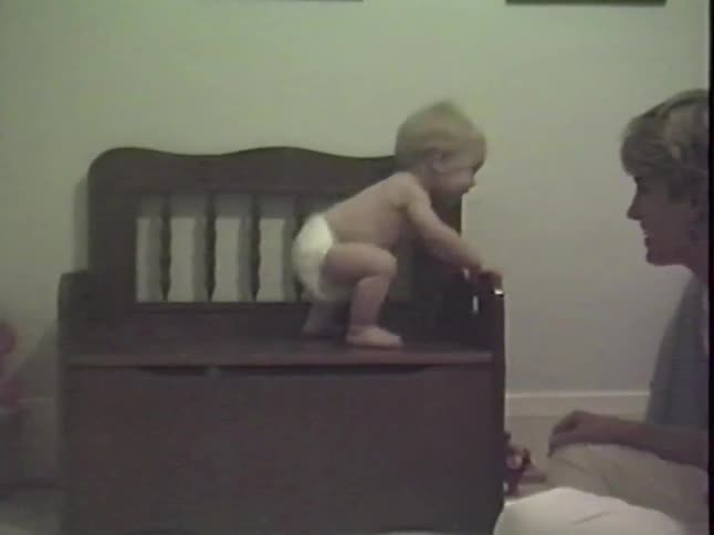 Vintage home video shows a baby in a diaper squatting and sitting on a toy box while their mother watches