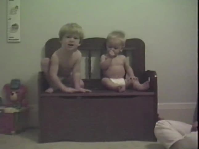 Vintage home video of a toddler and his baby brother sitting on a wooden toy chest in a bedroom