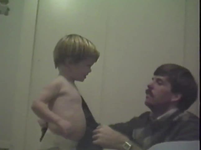 A father helps his toddler get dressed as the playful child raises his arms in the air in this vintage home video