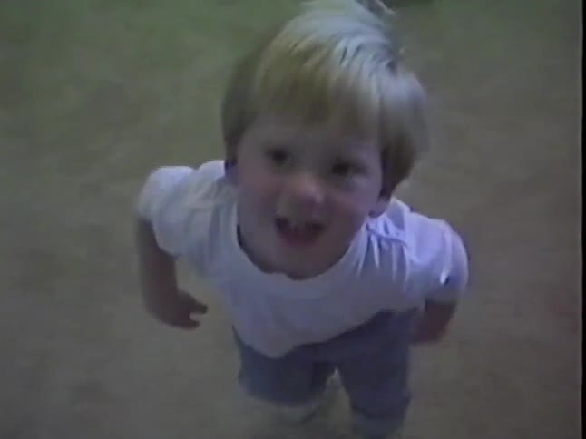 Vintage home video of a blond toddler looking up at the camera and walking down a carpeted hallway indoors