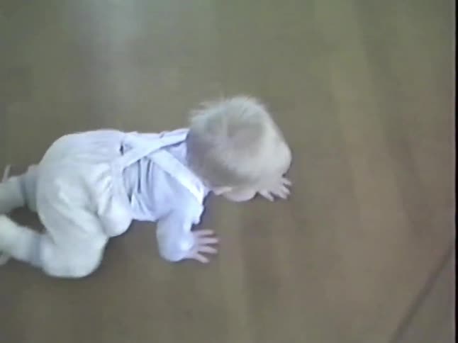 A baby in overalls crawls across a hardwood floor towards the feet of the person filming in this vintage home video