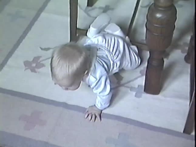A curious baby with big eyes crawls out from under a wooden chair in an authentic vintage home video