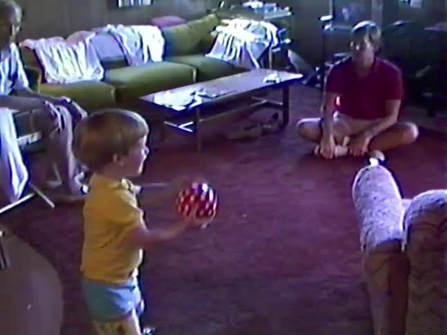 An adorable little boy plays catch with his father in the living room as his grandfather watches in this charming vintage home video