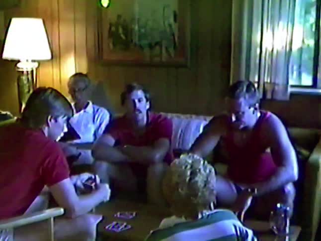 Vintage VHS footage shows a family playing a card game together in a cozy, wood-paneled living room