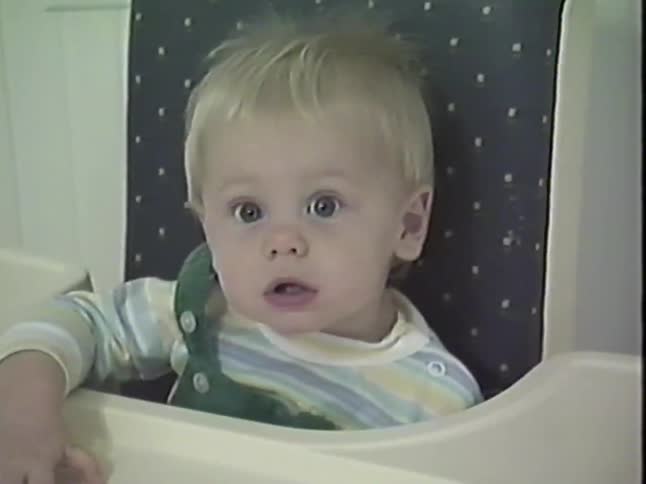 A cute blonde baby in a high chair makes funny faces and smiles at the camera in this vintage home video
