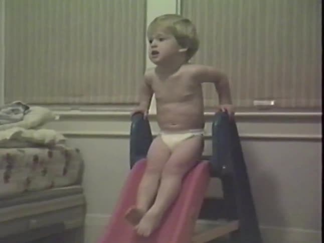 An adorable toddler in a diaper laughs with pure joy as he slowly slides down his toy slide in his bedroom