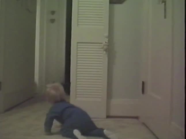 A vintage home video shows a toddler in a blue onesie crawling down a hallway after his father