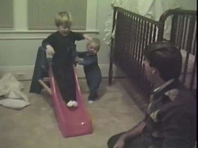 A young boy slides down a toy slide into his father's arms while his little brother watches in their bedroom