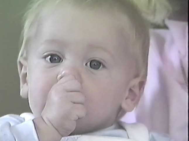 A sweet vintage home video shows a close-up of an adorable baby boy sucking his thumb as his smiling mother holds him