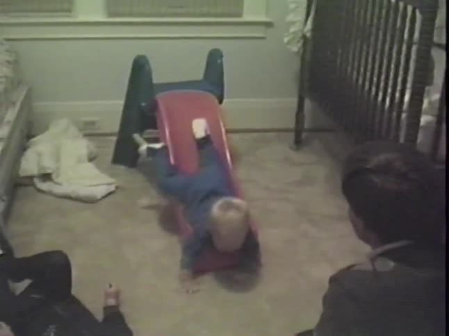 Vintage home video of a toddler in blue pajamas sliding headfirst down a toy slide as his father and sibling watch