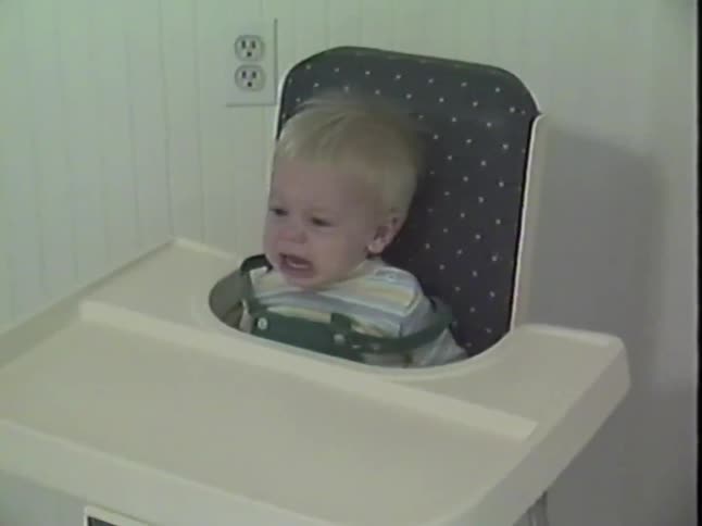 Vintage home video footage shows a cute blonde baby sitting in a high chair who begins to cry