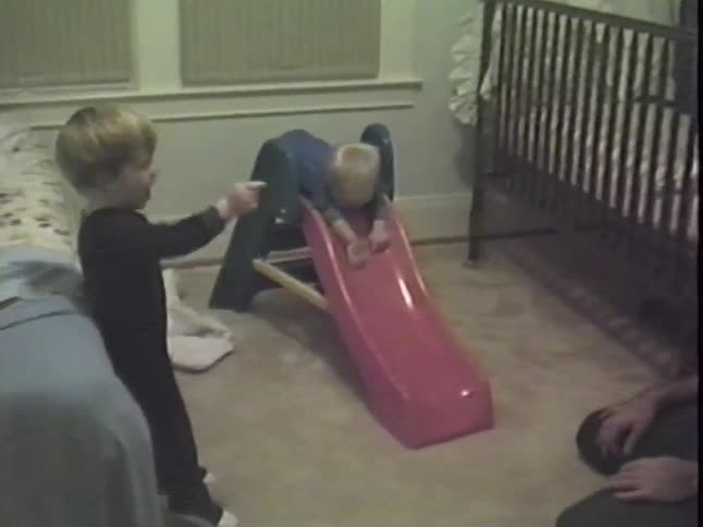 Vintage home video shows a cute toddler in a onesie sliding clumsily headfirst down a toy slide in his bedroom