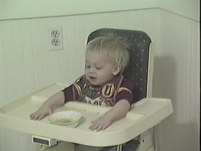 An adorable blond toddler in a highchair laughs hysterically, waves their arms, and points excitedly during mealtime in this vintage home video