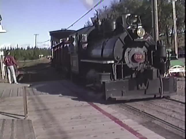 An old-fashioned steam locomotive pulls into a rustic train station on a sunny day, captured on vintage video