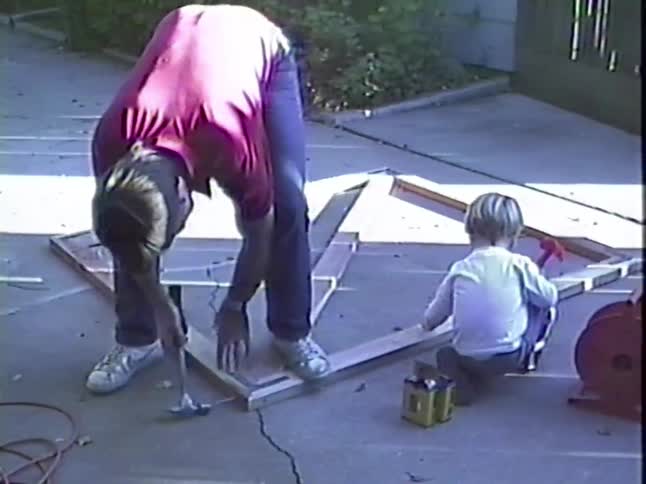 A father hammers a wooden frame on the driveway while his toddler son 'helps' with a toy hammer in this heartwarming vintage home video