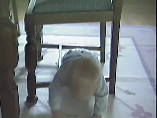 An adorable baby in overalls smiles from under a chair before crawling across the floor in this heartwarming vintage home video