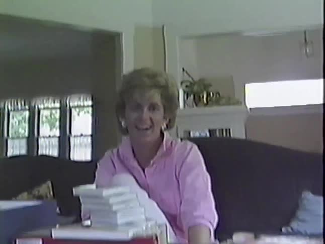 A vintage home video captures a woman with 80s style laughing and talking while sitting on a couch in her living room