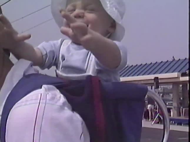 A smiling baby in a sun hat laughs while playing with a blue balloon from a backpack carrier on his father's shoulders