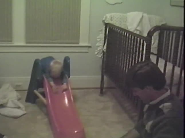 A happy toddler claps with excitement before sliding headfirst down a small slide into his father's arms in his bedroom