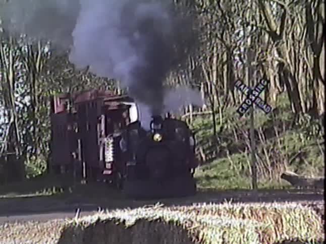 A vintage steam train billows thick smoke as it slowly approaches a rural railroad crossing in the woods