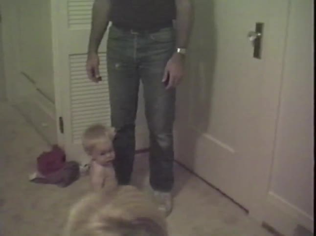 A cute blonde baby in a diaper crawls on a carpeted floor, hugs their father's leg, then crawls towards the camera in a vintage VHS home video
