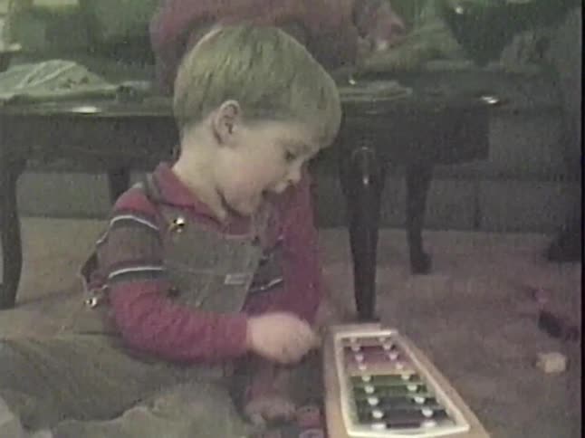 A vintage home movie shows a young boy in overalls playing with a toy xylophone on the living room floor