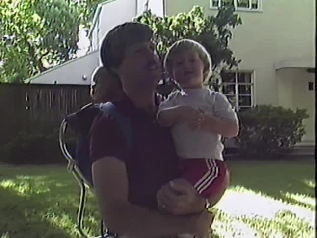 A loving father holds his toddler, giving them a kiss, while another baby rides along in a backpack carrier in their sunny backyard
