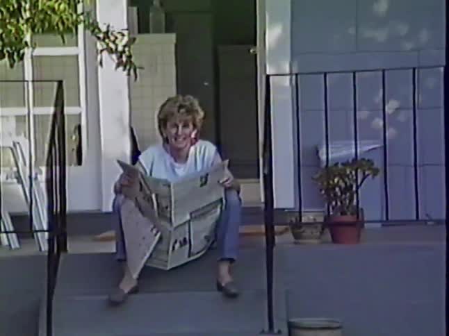 A woman reads the newspaper on her front porch steps before smiling and waving at the camera in this vintage home video