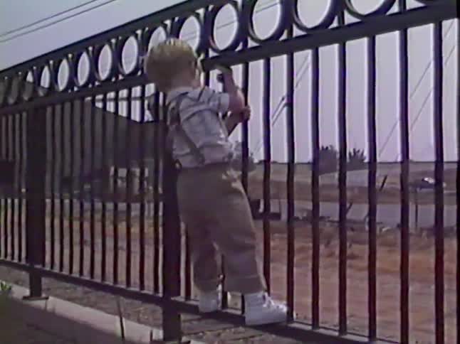 A curious toddler in suspenders playfully climbs on a metal fence overlooking a rustic area with railroad tracks