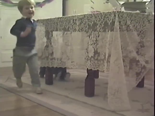 A happy toddler crawls under a table with a lace tablecloth while playing a game of peek-a-boo with his mother