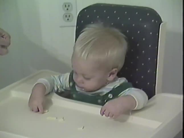 A cute blond baby wearing green overalls sits in a high chair and happily eats snacks from the tray