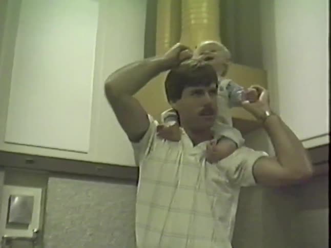 Authentic vintage home video shows a happy father with a mustache giving his laughing baby boy a shoulder ride in the kitchen