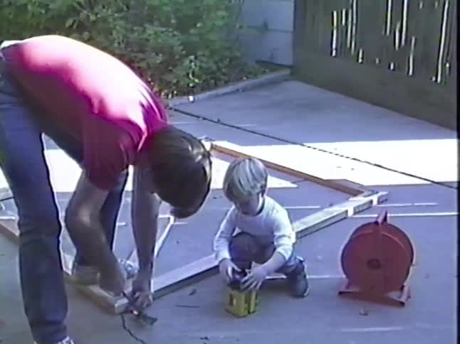 Vintage footage shows a father working on a DIY project in the driveway while his young son watches and plays nearby