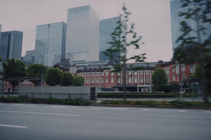 A vehicle's point of view driving past the historic Tokyo Station, with modern skyscrapers rising behind it in the city center