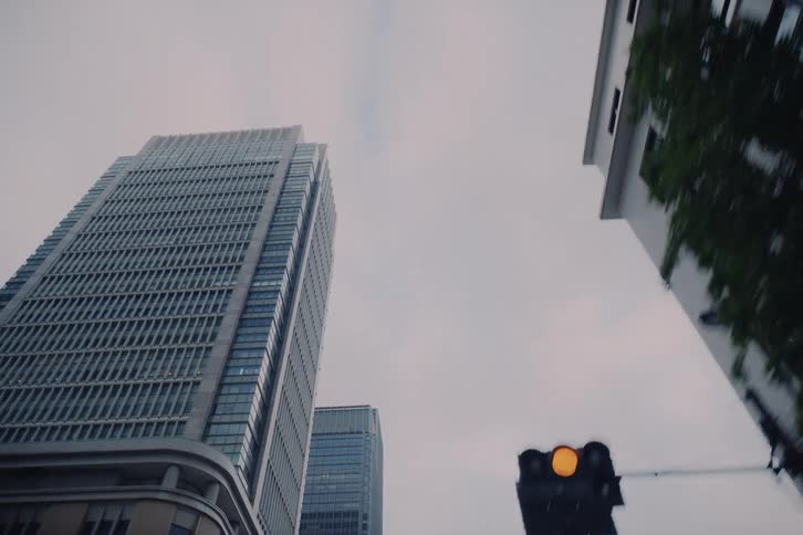 A low-angle, upward-panning view from a moving vehicle captures the towering skyscrapers of a modern city against an overcast sky
