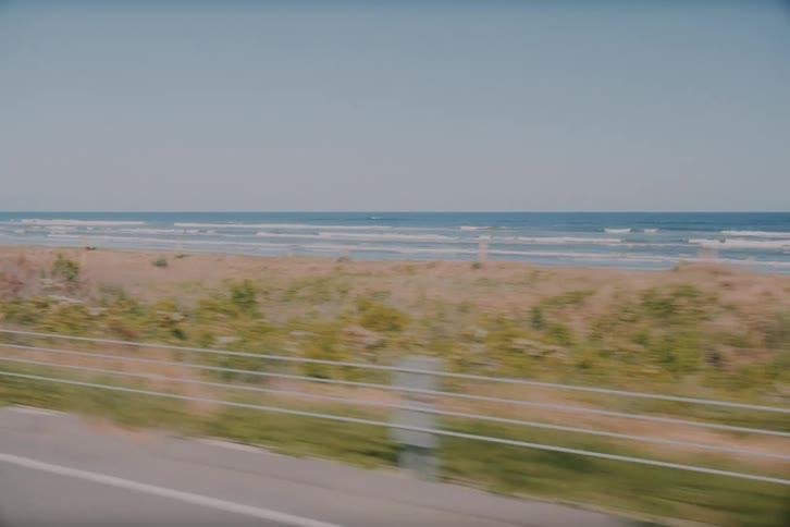 A scenic view from a moving vehicle driving along a coastal road, with the sandy beach and ocean waves in the background