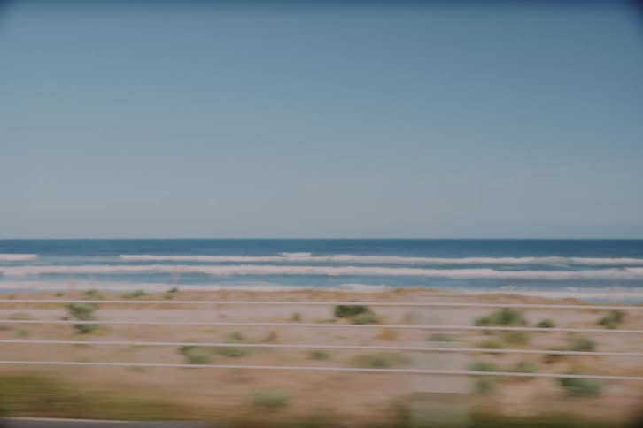 A blurry, nostalgic view from a moving car window shows the ocean waves crashing on a sandy beach on a sunny day