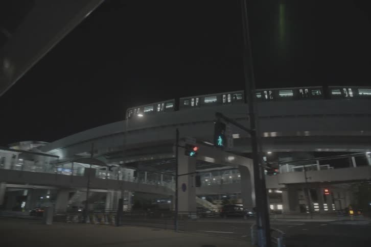 A monorail train passes over a deserted city street intersection at night as the camera pans left