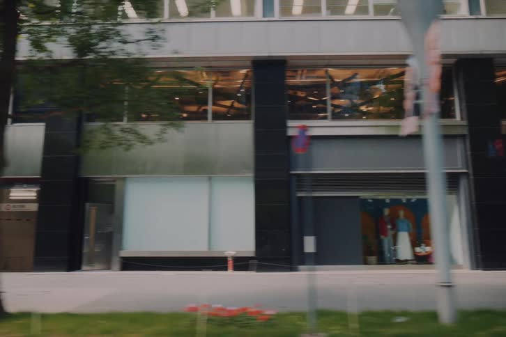 A smooth tracking shot captures pedestrians walking along a tree-lined street past modern storefronts in a Japanese city