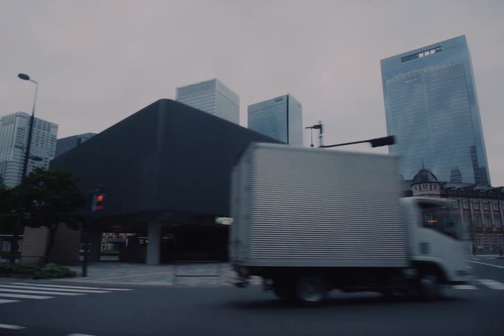 A tracking shot from a moving vehicle captures the historic Tokyo Station building set against the modern skyscraper skyline on an overcast day