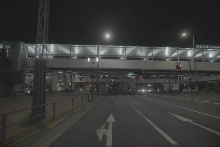 A slow-motion point-of-view shot moving towards the illuminated Toyosu Market overpass on a quiet city street in Tokyo at night