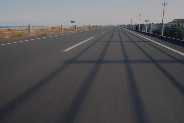 A low-angle point-of-view shot driving along a straight coastal highway, with long shadows from the guardrail stretching across the asphalt
