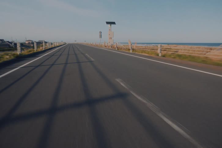 Point-of-view footage driving along an empty, straight coastal road with the ocean on one side during a sunny day