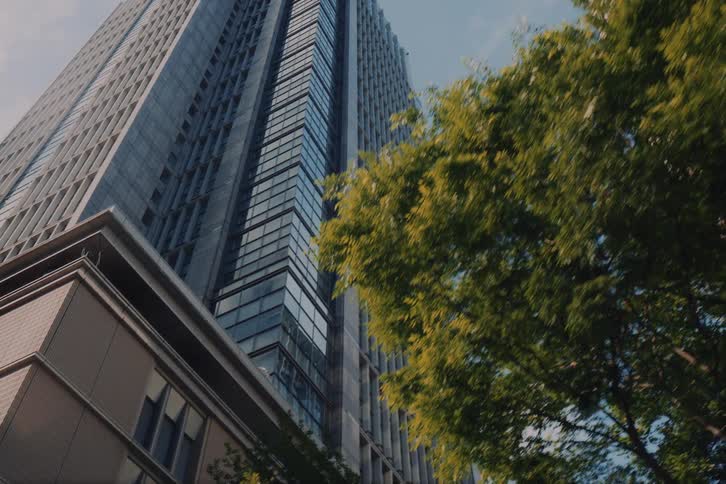 Driving through a city looking up at modern glass skyscrapers framed by vibrant green trees on a sunny day