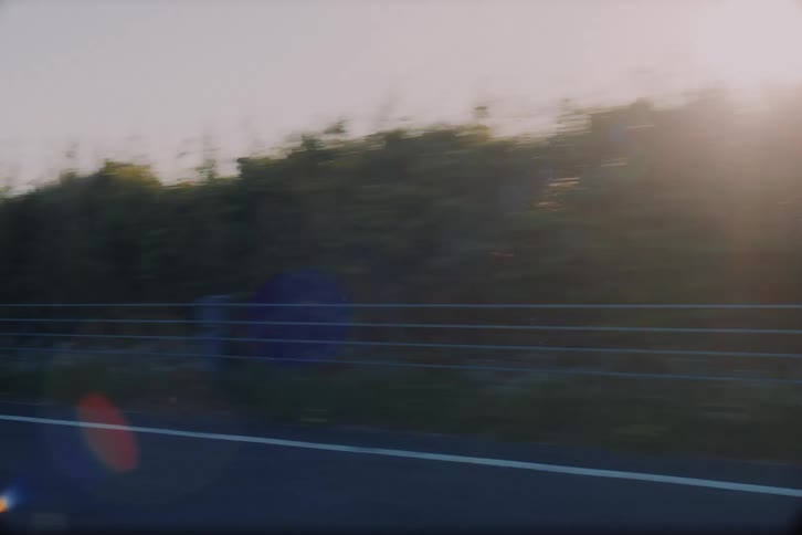 A dreamy, sun-flared side view from a car transitions to a point-of-view shot driving down an empty rural highway at sunrise