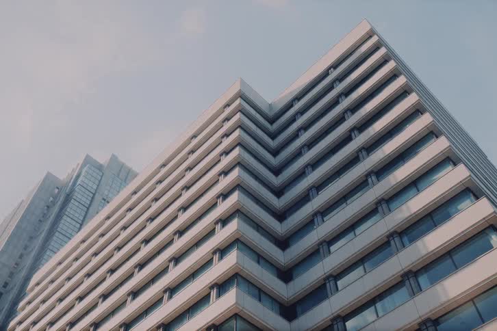 Driving past a contemporary city building, a low-angle tracking shot captures the architectural details as the scene transitions from bright daylight to evening